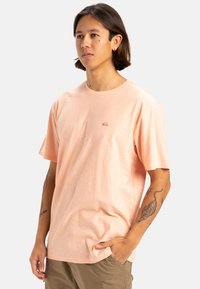 Camiseta corta de manga con cuello redondo en color melocotón claro, hecha de un tejido texturizado, con un pequeño logo en el pecho. Combinada con pantalones cortos caqui.