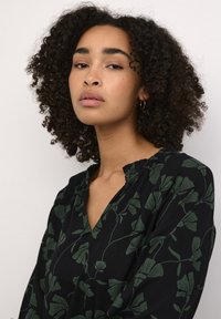 Blouse noire à décolleté en V profond, manches longues et motif floral vert. Tissu doux avec une texture lisse.