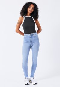 Bodysuit negro sin mangas con detalles blancos, jeans ajustados de cintura alta en azul claro y zapatillas deportivas blancas. Fondo liso.