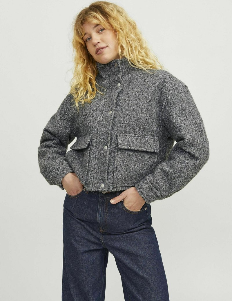 Giacca grigia corta realizzata in tessuto texture, caratterizzata da un collo alto, chiusure a bottoni e due tasche frontali. Abbinata a jeans blu scuri.