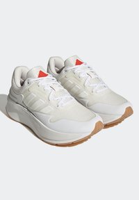 Chaussures de sport blanches avec une tige en mesh, des panneaux texturés et une semelle en caoutchouc beige. Elles présentent des accents rouges sur la languette et trois bandes latérales.