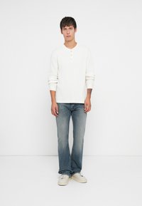 Chemise henley blanche à manches longues avec trois boutons, associée à un jean flare bleu clair et des baskets blanches, sur un fond uni.