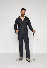 Mörkblå utomhusjacka med huva och dragkedja. Matchas med byxor i samma färg och ljusa vandringskängor. Använder vandringsstavar.