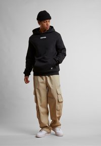Schwarzer Kapuzenpullover mit einem Logo, kombiniert mit beigen Cargo-Hosen, die mehrere Taschen haben. Weiße Sneakers runden das Outfit ab.
