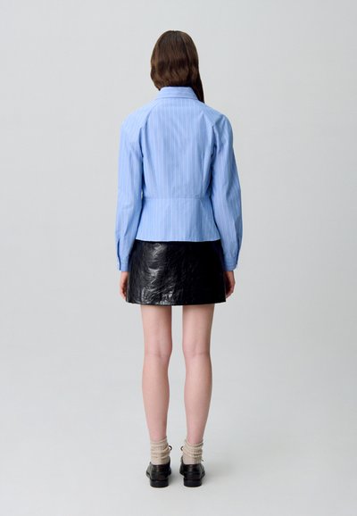 Camisa de botones a rayas de color azul claro, con mangas largas, combinada con una mini falda de cuero negra. El cabello es largo y sin adornos.