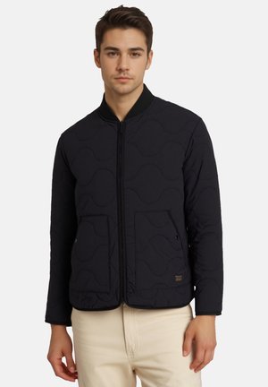 Ung mand iført en sort quiltet jakke med lynlås og to forlommer samt beige bukser mod en ensfarvet baggrund.