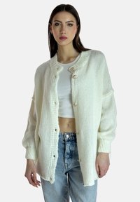 Weißer gestrickter Cardigan mit Rundhalsausschnitt, ausgestattet mit vier Vorderknöpfen, lockeren Ärmeln und einem strukturierten Muster. Kombiniert mit hochtaillierten Jeans.