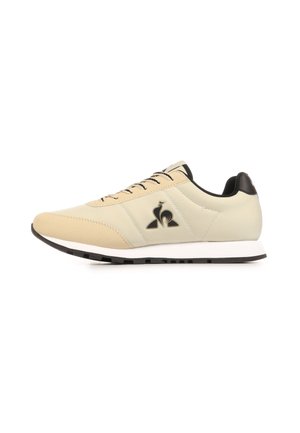 Beige-cremefarbener Sneaker mit niedrigem Schaft, schwarzem Logo, schwarzen Schnürsenkeln und weißer Zwischensohle auf weißem Hintergrund, Seitenansicht.