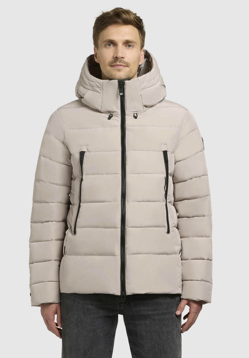 Lys beige pufferjakke med hætte, der har sorte lynlåse, rektangulært quiltet mønster og to lynlåslommer foran.