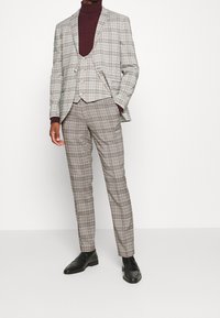 Karoanzug in Grau, Braun und Schwarz mit maßgeschneiderter Passform, kombiniert mit einem burgunderfarbenen Rollkragenpullover. Mit einem Zwei-Knopf-Jacket und eleganten Schuhen.