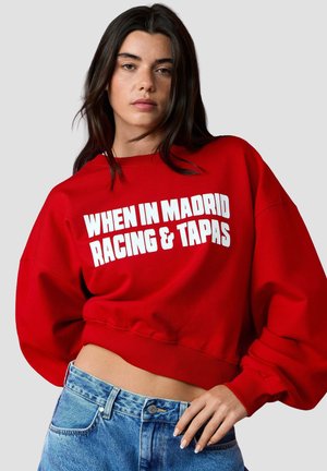 Jonge vrouw met lang donker haar die een rode cropped sweater draagt met witte tekst "WHEN IN MADRID RACING & TAPAS" en een blauwe spijkerbroek.