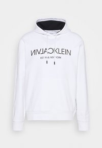 Valkoinen huppari, joka on valmistettu puuvillasekoitteesta. Siinä on edessä kengurupocket, nauhakuoma ja musta logo-teksti painettuna eteen.