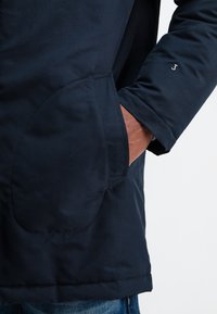 Marineblauwe jas met een gladde textuur, voorzien van een groot opgestikt zak, gestikte accenten en een klein logo op de mouwboord.