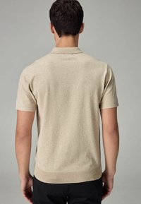 Beige lyhythihainen polo-paita, jossa on litteä neulekaulus, ribbineulereuna ja sileä pinta. Ei näkyviä kuvioita tai yksityiskohtia.