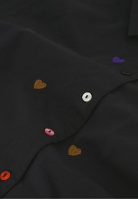 Zwarte stoffen shirt met kleine geborduurde hartjes in oranje, paars en rood. Vastgemaakt met een mix van gekleurde knopen.