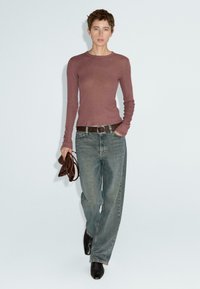 Langärmliges bordeauxrotes Oberteil kombiniert mit ausgebleichten blauen Jeans. Brauner Gürtel und schwarze hohe Stiefel. Die Person hält eine braune Handtasche. Einfacher Hintergrund.