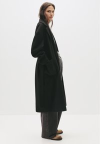 Čierny dlhý kabát s golierom, bočnými vreckami a jednoduchým dizajnom, preložený cez šedé oblečenie, v kombinácii s voľnými tmavými džínsy a svetlými topánkami.