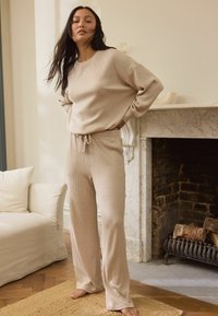 Beige ribbed knit loungewear setti, joka sisältää rento-mallisen paidan pitkillä hihoilla ja nauhalla säädettävät housut. Pehmeä materiaali ja rento muotoilu.