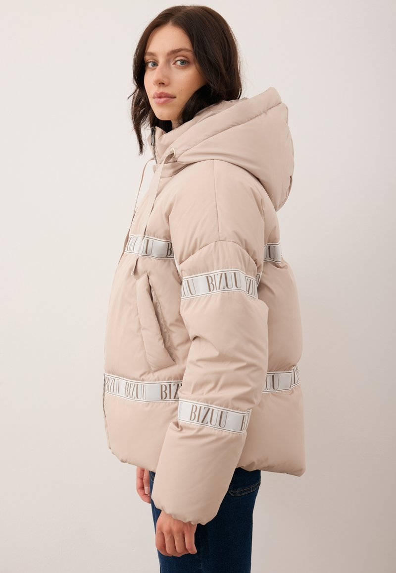 Kobieta w beżowej kurtce ocieplanej z kapturem i białymi paskami z logo "BIZUU", stojąca w profilu na tle jasnej ściany.