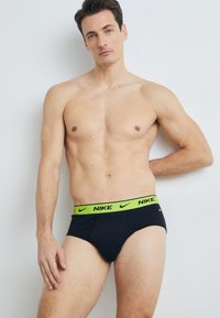 Schwarze Unterwäsche mit einem leuchtend grünen Nike-Logo-Bund, ausgestattet mit einer engen Passform und weichem Stoff. Das Model steht vor einem neutralen Hintergrund.