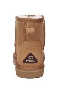 Tan suede ankle boot with a rounded toe, soft fur lining, and a branded brown leather patch on the heel. Features textured stitching.