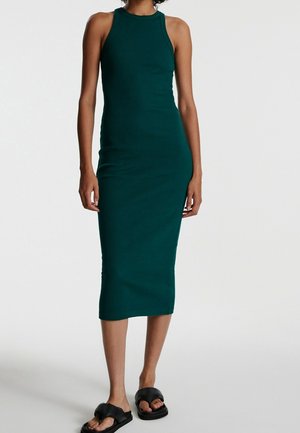 Femme portant une robe midi ajustée sans manches de couleur vert foncé associée à des sandales noires à bouts ouverts, debout devant un fond blanc uni.