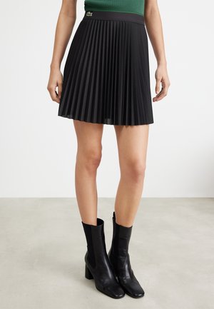Zwarte plissé rok met een aansluitende tailleband en Lacoste-logo, gecombineerd met zwarte leren enkellaarsjes op een neutrale, gestructureerde vloer.