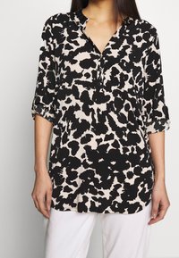 Svart och beige blus med ett leopardmönster, med knäppning i V-hals, trekvartslånga ärmar och en slät tygtextur.