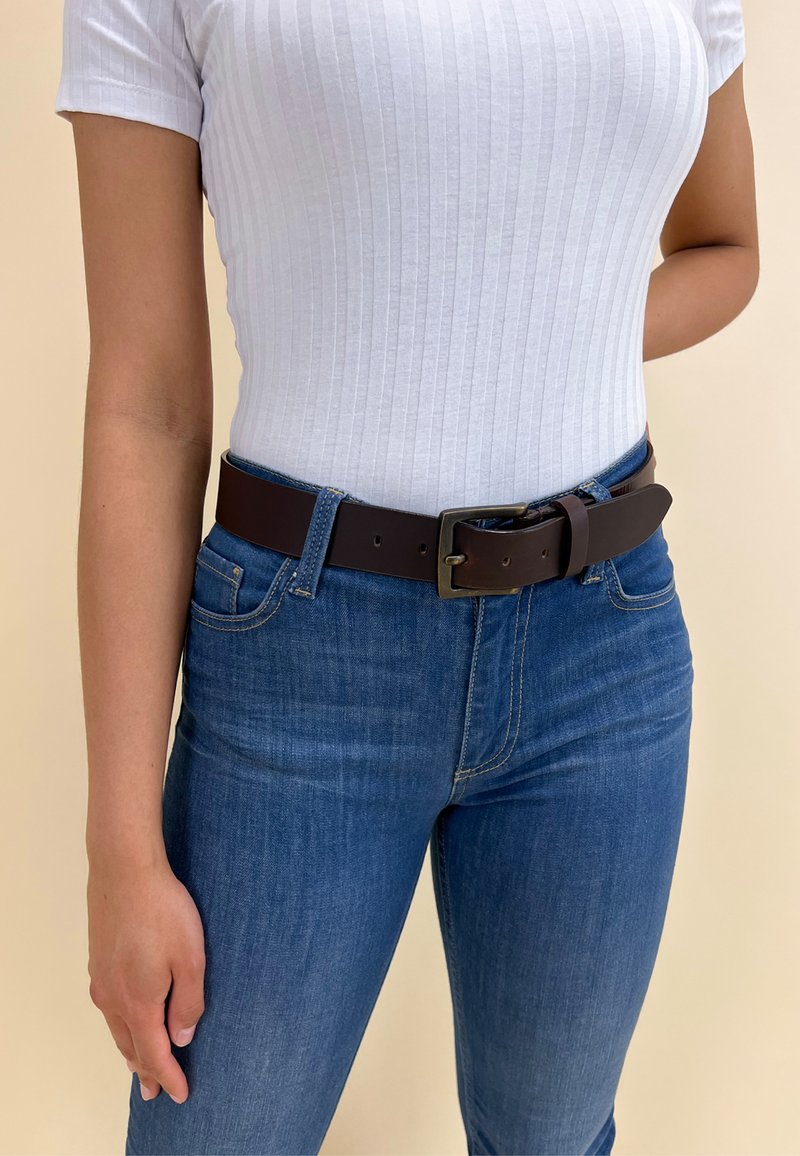 Brun læderbælte med en sølv rektangulær spænde, båret over en hvid ribbet top og blå denim jeans. Glat tekstur, afslappet pasform.