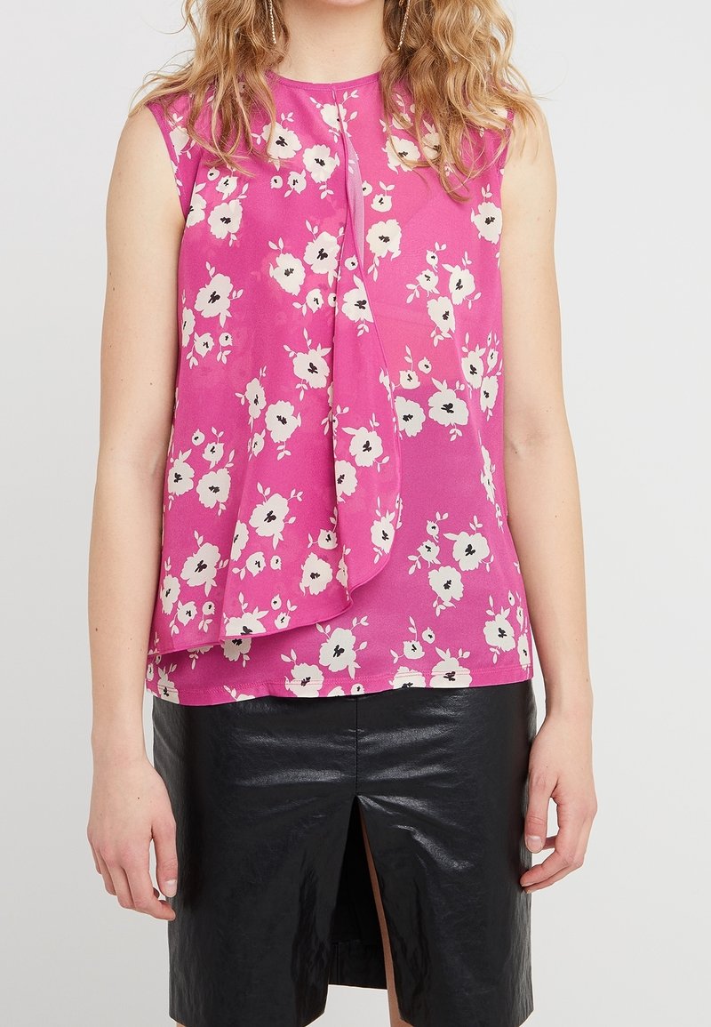 Haut sans manches rose à motifs floraux avec un devant drapé, présentant des motifs de fleurs blanches et noires ; associé à une jupe noire en faux cuir brillant.