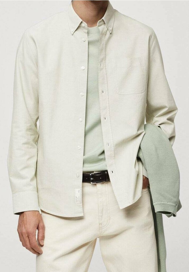 Homme portant une chemise beige clair boutonnée sur un t-shirt vert pâle et un pantalon beige, tenant un pull en maille vert plié dans une main.
