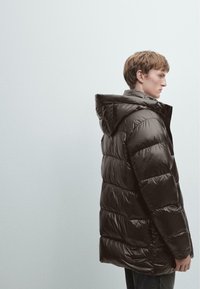 Cappotto imbottito marrone scuro, con cappuccio, texture lucida, design trapuntato e chiusura frontale con zip. Indossato sopra una camicia chiara e jeans scuri.