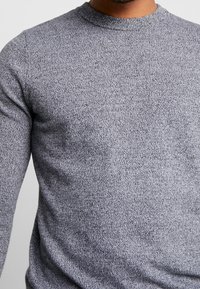 Szary sweter dzianinowy z okrągłym dekoltem, z teksturowanym wzorem i długimi rękawami. Materiał wydaje się miękki i elastyczny.