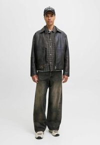 Een jonge man staat zelfverzekerd in een zwart leren jack, een geruit overhemd, brede spijkerbroeken en sneakers tegen een neutrale achtergrond.