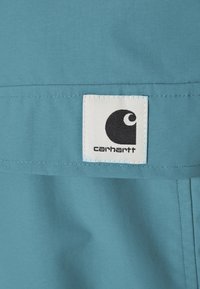 Close-up van een teal stoffen kledingstuk met een vierkant label met een zwart logo en een witte achtergrond, genaaid aan de linkerkant. Gladde textuur.