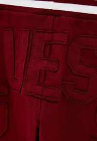 Burgundin väriset jogger-housut, joissa on tekstuurilla ommeltu "GUESS"-merkintä. Ominaisuuksiin kuuluu vaalean valkoinen vyötärönauha ja sileä, pehmeä kangas.