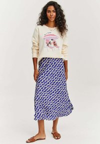 Krémová mikina s grafickým potiskem, spárovaná s modrou sukní s bílými vlnkovými vzory. Doplňuje ji obuv na plochém podpatku hnědé barvy.