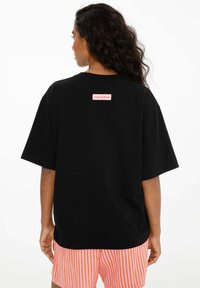 Žena s dlouhými kudrnatými vlasy, oblečená v volném černém tričku s nápisem "Club Tropicana" na zadní straně a oranžovo-bílých pruhovaných šortkách.