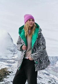 Žena v ružovej čiapke, náražovej páperovej bunde so zebrovaným vzorom a zeleným podšívkou, béžovom svetri a čiernych nohaviciach na zasneženom horskom pozadí.