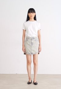 Witte katoenen t-shirt met een klein logo, gecombineerd met een lichte denim minirok met knoopsluiting en voorzakken. Zwarte platte schoenen.