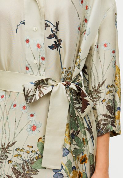 Haut style kimono à imprimé floral en tissu beige clair, présentant des motifs botaniques en vert, bleu et rouge avec un détail ceinturé à la taille.