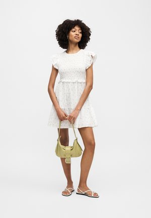 Γυναίκα με κοντό λευκό φόρεμα με floral σχέδιο και μανίκια με βολάν, κρατώντας μια μικρή ανοιχτοπράσινη τσάντα, φοράει λευκά πέδιλα με λουριά, στέκεται μπροστά σε λευκό φόντο.