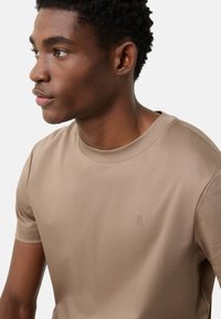 Krótki rękaw, jasnobrązowa koszulka z gładkiego materiału z okrągłym dekoltem i subtelnym logo "B" po lewej stronie. Prosty, czysty design.