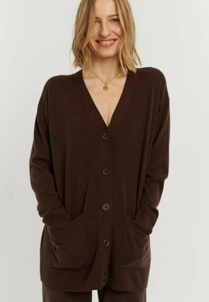 Donna sorridente che indossa un cardigan oversize marrone scuro con bottoni e tasche frontali e una collana d'oro su uno sfondo semplice.