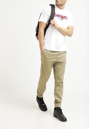 Man met wit T-shirt met opdruk, beige joggerbroek, zwarte sneakers en zwarte rugzak, lopend met één hand in zijn zak.