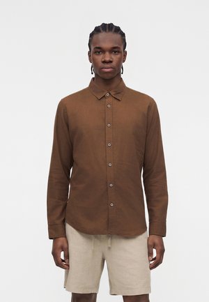 Νέος άνδρας με πλεξίδες στα μαλλιά, φορώντας καφέ πουκάμισο με κουμπιά και μπεζ σορτς, στέκεται μπροστά σε απλό λευκό φόντο.