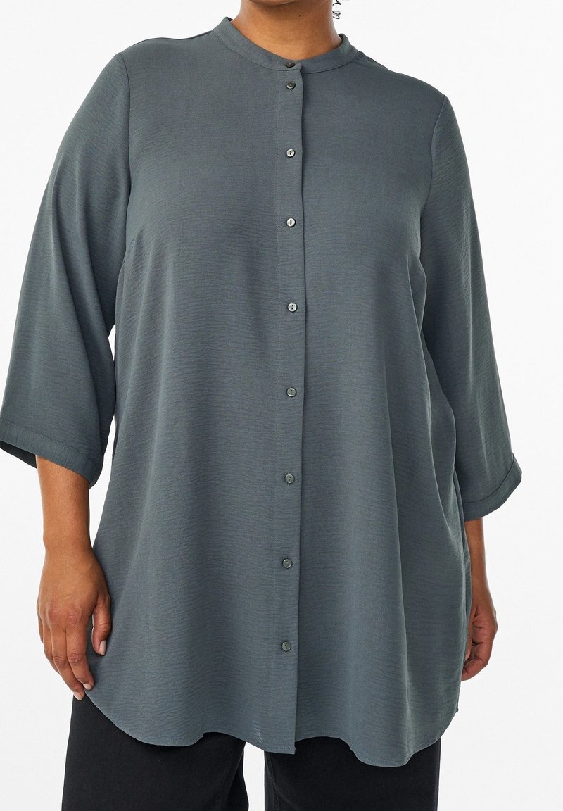 Femme portant une tunique ample, boutonnée, gris foncé, à manches trois-quarts et col rond, associée à un pantalon noir.
