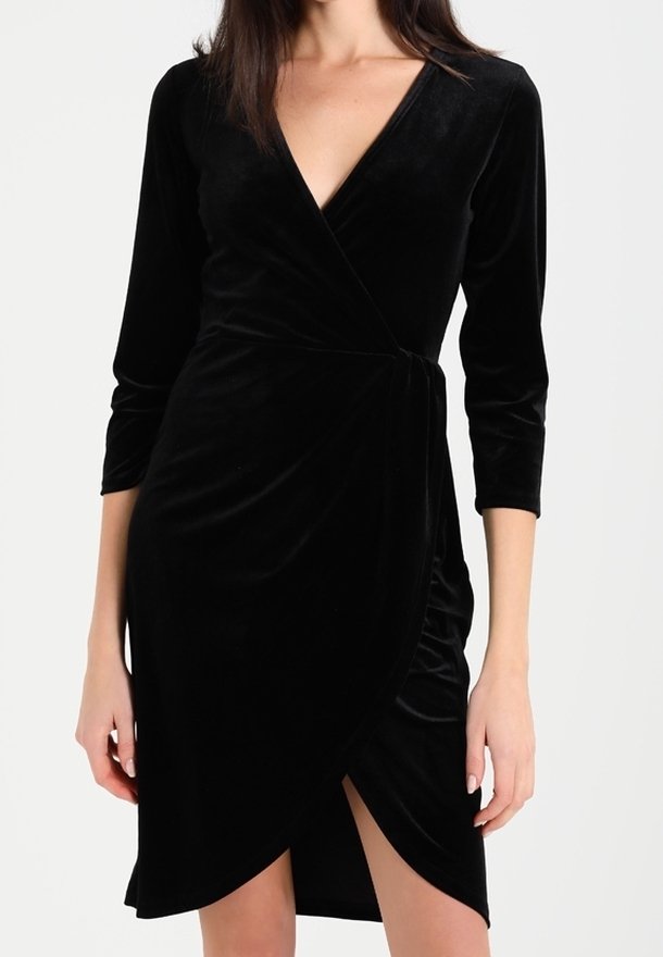 Femme portant une robe portefeuille en velours noire avec des manches trois-quarts et un décolleté en V, avec un ourlet tulipe à l'avant.