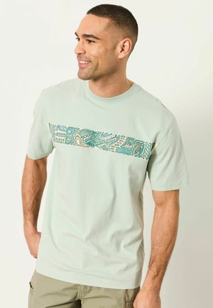 Mann mit kurzen Haaren trägt ein hellgrünes T-Shirt mit abstraktem, horizontalem Streifenmuster, beige Cargoshorts, lächelt und blickt nach links.