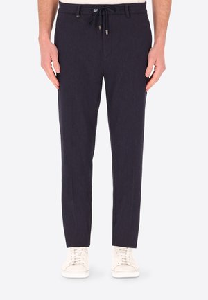 Uomo che indossa pantaloni su misura blu navy con vita con coulisse e sneakers bianche, in piedi davanti a uno sfondo uniforme.